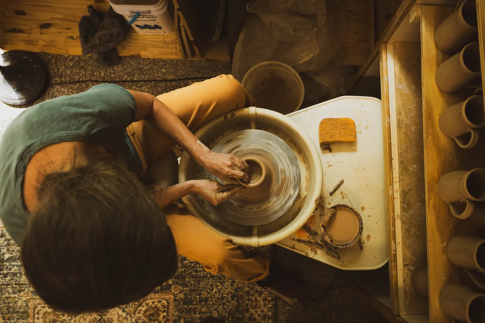Overhead view of Karissa Masse centering clay on the pottery wheel, with a shelf of mugs beside her.