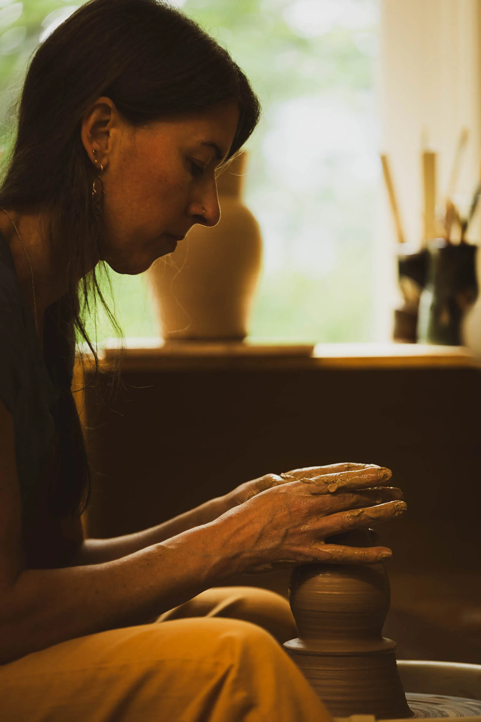 Karissa Masse in profile, shaping a vase on the wheel with clay-covered hands beside a sunlit window.