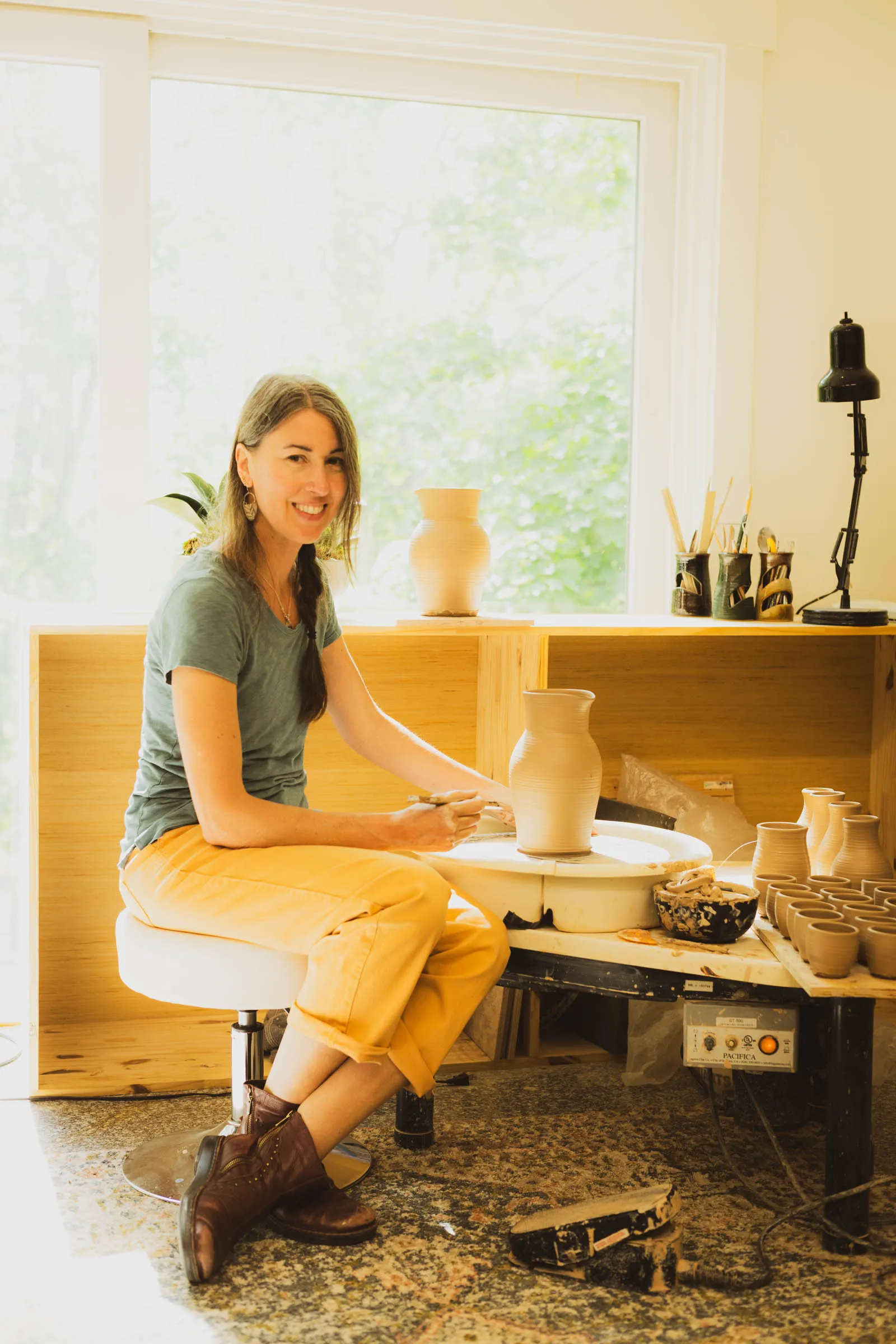 Karissa Masse smiling at her pottery wheel in a sunlit studio, with a tall unglazed jug in progress and a row of mugs nearby.