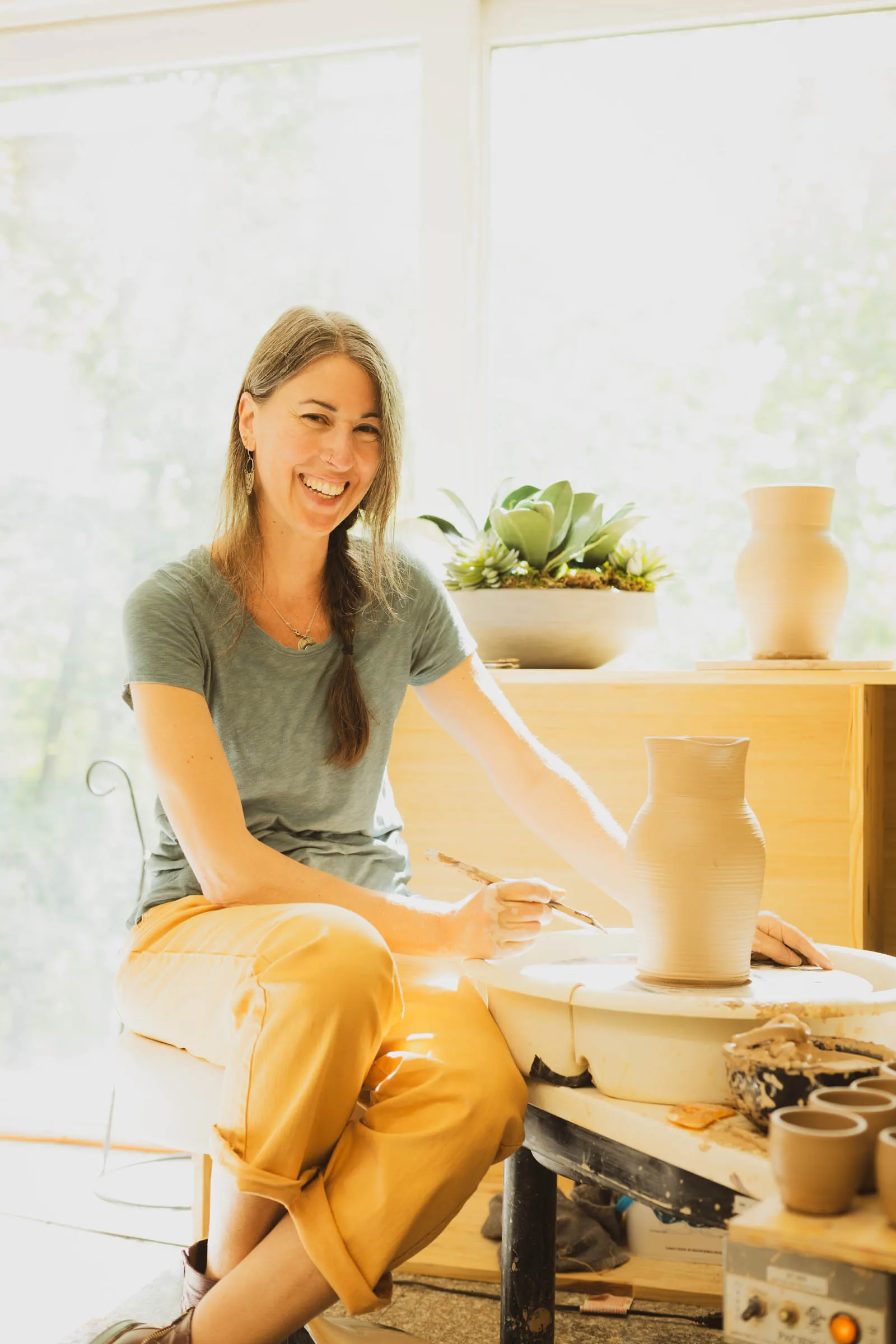 Karissa Masse laughing as she works on a clay pitcher on the wheel in a bright, sunlit studio.