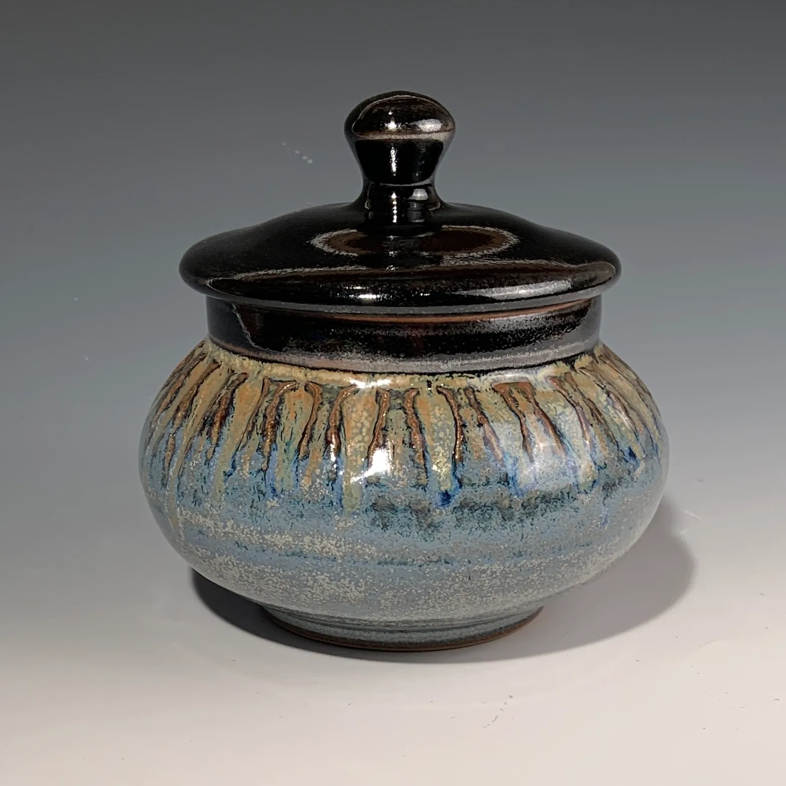 Peacock lidded jar with a glossy dark tenmoku lid and a blue glaze banded with amber drips.
