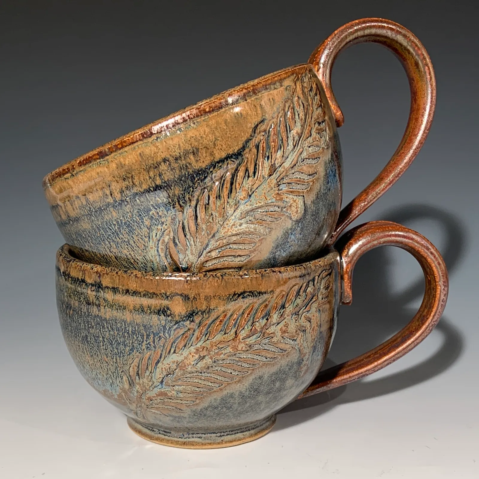 Two stacked Peacock cups with carved feather impressions in a blue and amber glaze, with copper-toned handles.