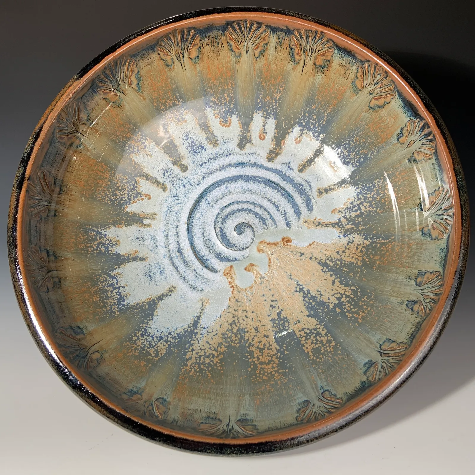 Peacock bowl seen from above, with a white and blue swirl at the center, carved feather designs around the rim and a dark tenmoku edge.