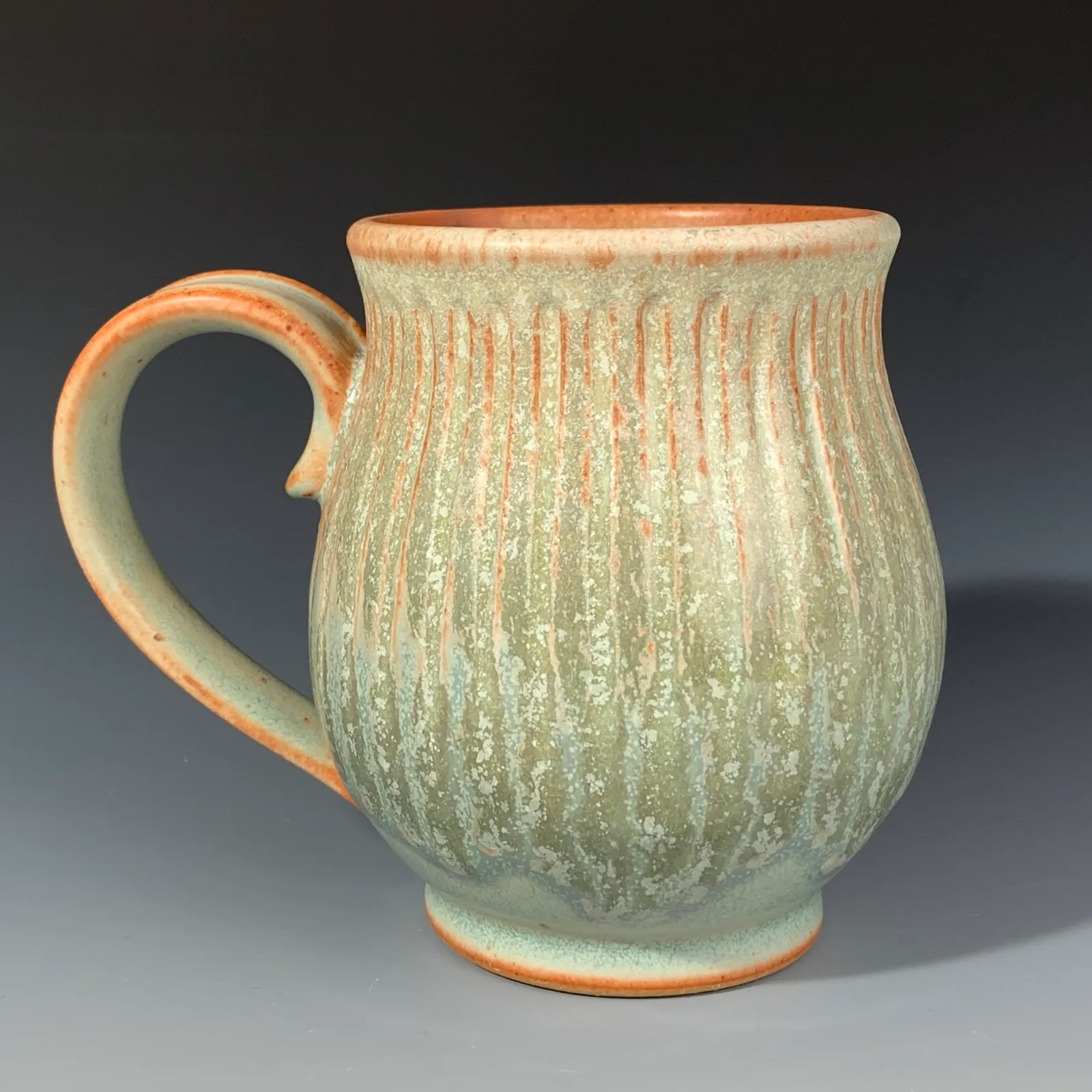 Willow mug in pale green glaze with carved vertical lines, an orange-brown rim and a curled handle.
