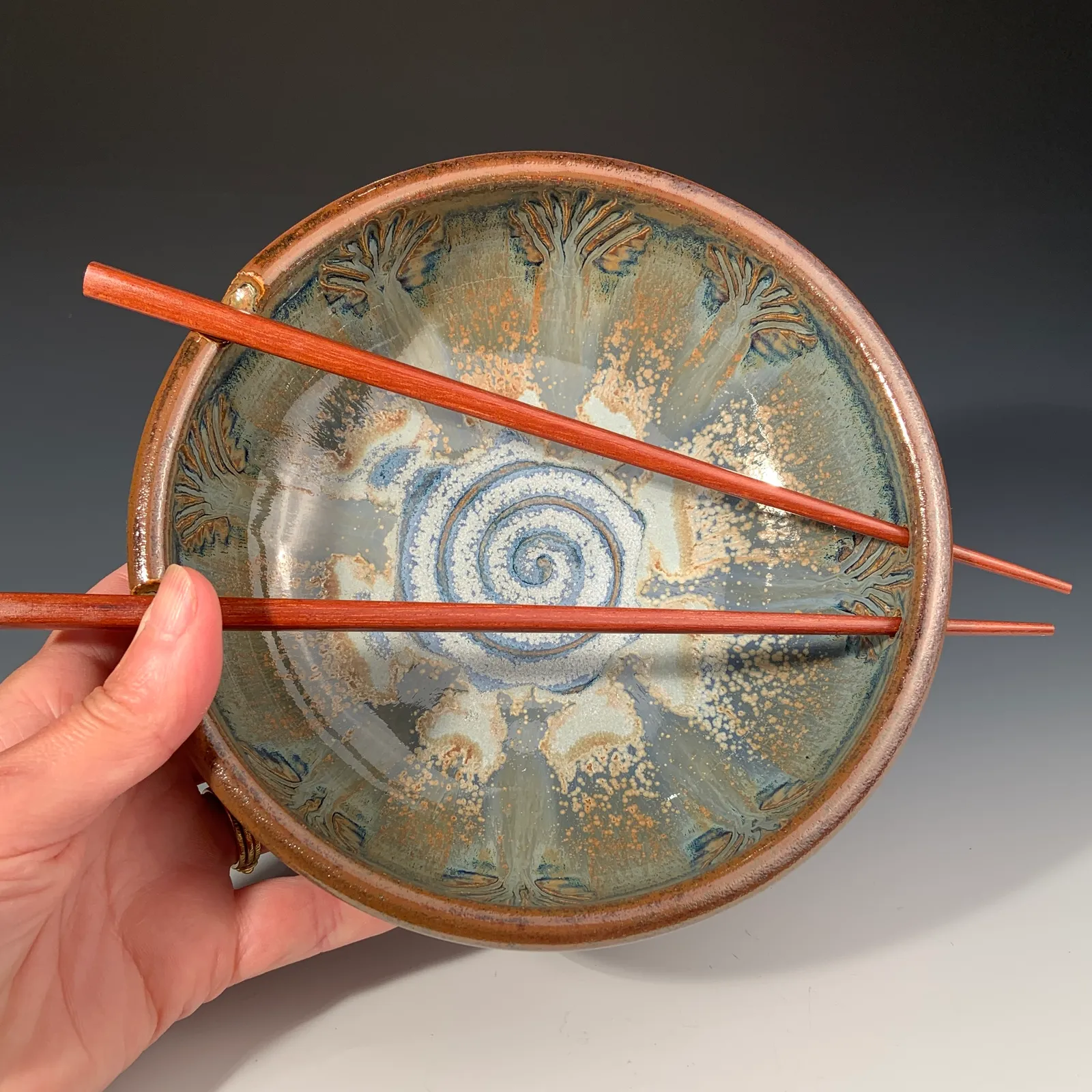 Peacock bowl held in one hand with red wooden chopsticks resting across it, showing a blue and white spiral center and carved feather designs.